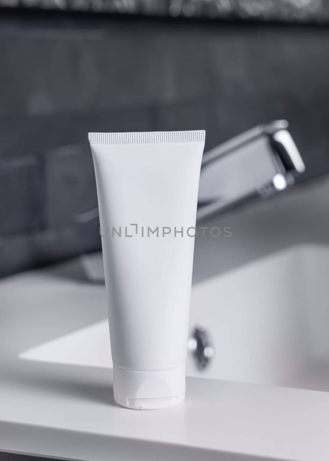 White cream squeeze tube near modern faucet on basin in dark grey masculine bathroom close up, packaging mockup. Natural unbranded cosmetics with copy space. Spa and wellness concept