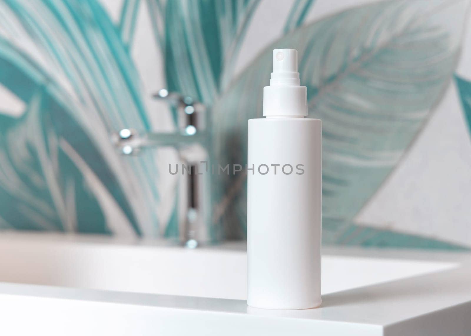 White cosmetic spray bottle on washbasin with faucet in green tropical bathroom close up, packaging mockup. Natural plant unbranded lotion or toner container, copy space. Spa and wellness concept