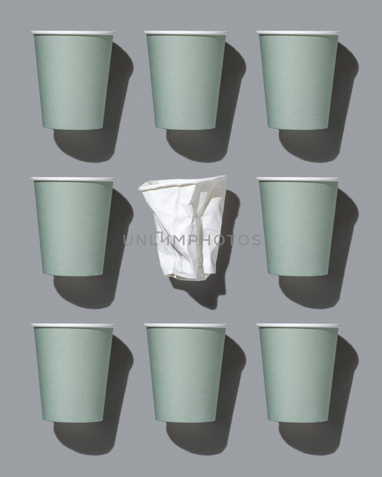 Disposable new light green paper cups and crumpled used white one on gray top view, pattern, hard shadow, mockup with copy space. Empty cardboard coffee cups flat lay. Consumerism concept