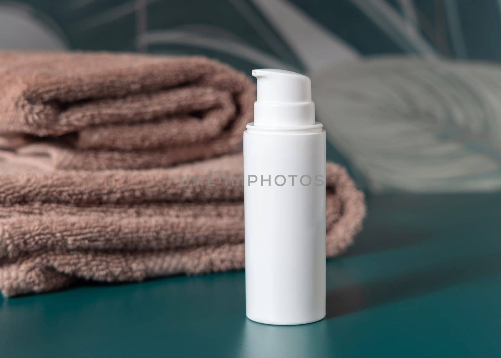 White cream one pump bottle near brown folded towels in dark green tropical bathroom close up, packaging mockup. Natural plant unbranded cosmetic container, copy space. Spa and wellness concept