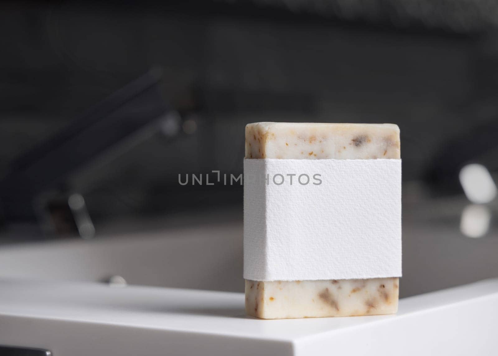 Beige handmade soap bar with blank label on washbasin near faucet in modern dark grey masculine bathroom close up, mockup. Natural herbal product for daily male hygiene and bodycare