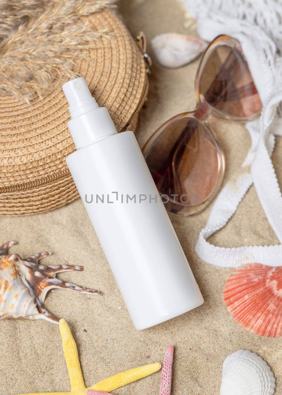 White blank spray bottle, sunglasses, towel, starfishes and shells on a sandy beach, top view. Sunscreen cosmetics mockup. Summer vacation and spf uv-protecting skin care concept