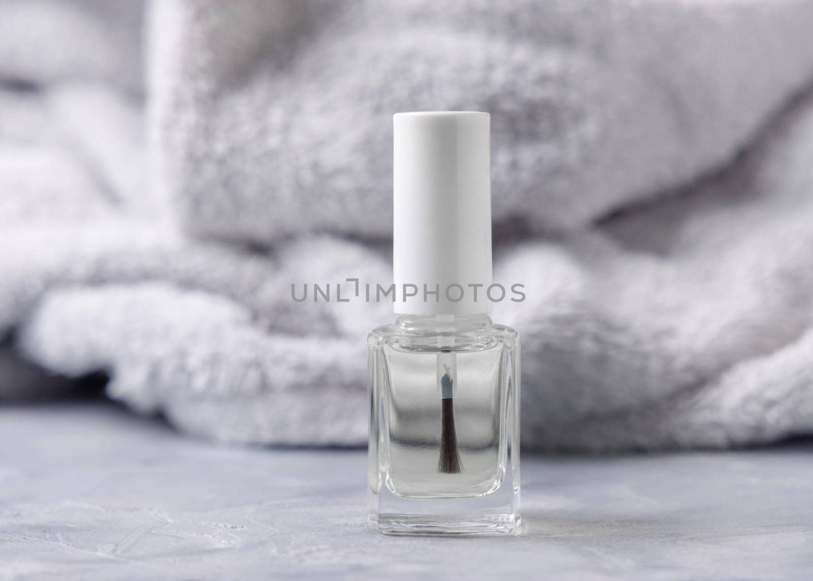 Clear glass refillable bottle with white blank brush cap near grey bath towel on grey close up, cosmetics mockup. Natural skincare nail product, minimal aesthetic