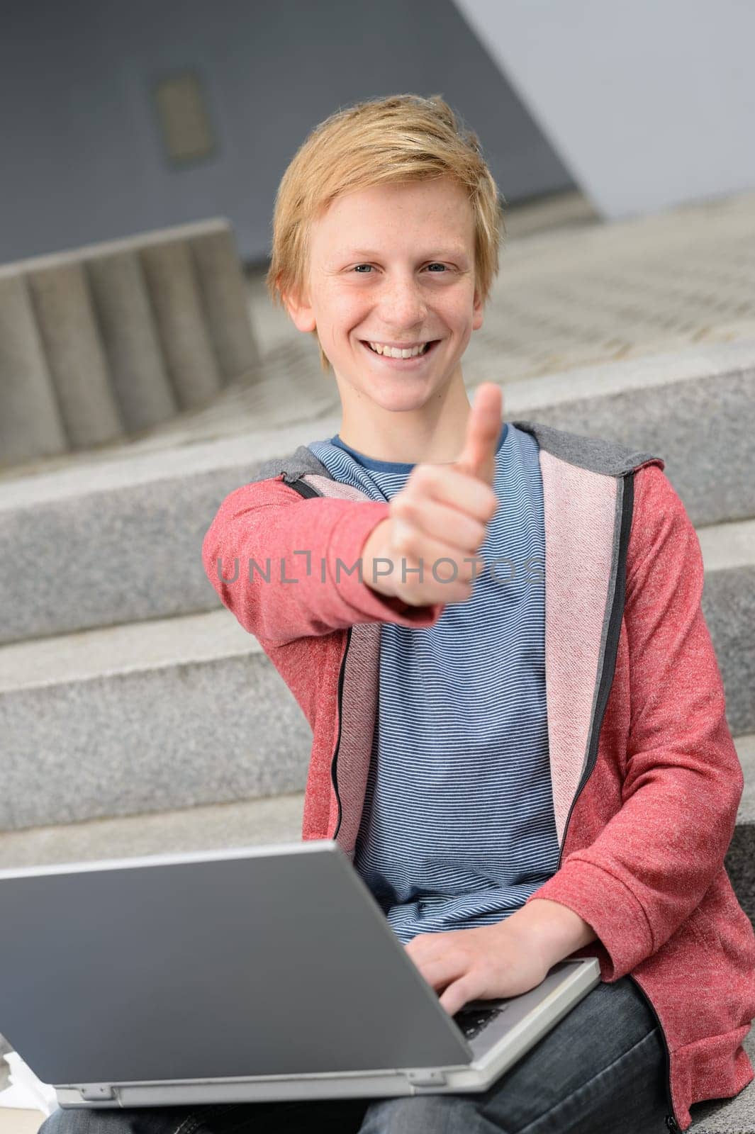 Successful teenage student show thumb-up  sitting with laptop on steps