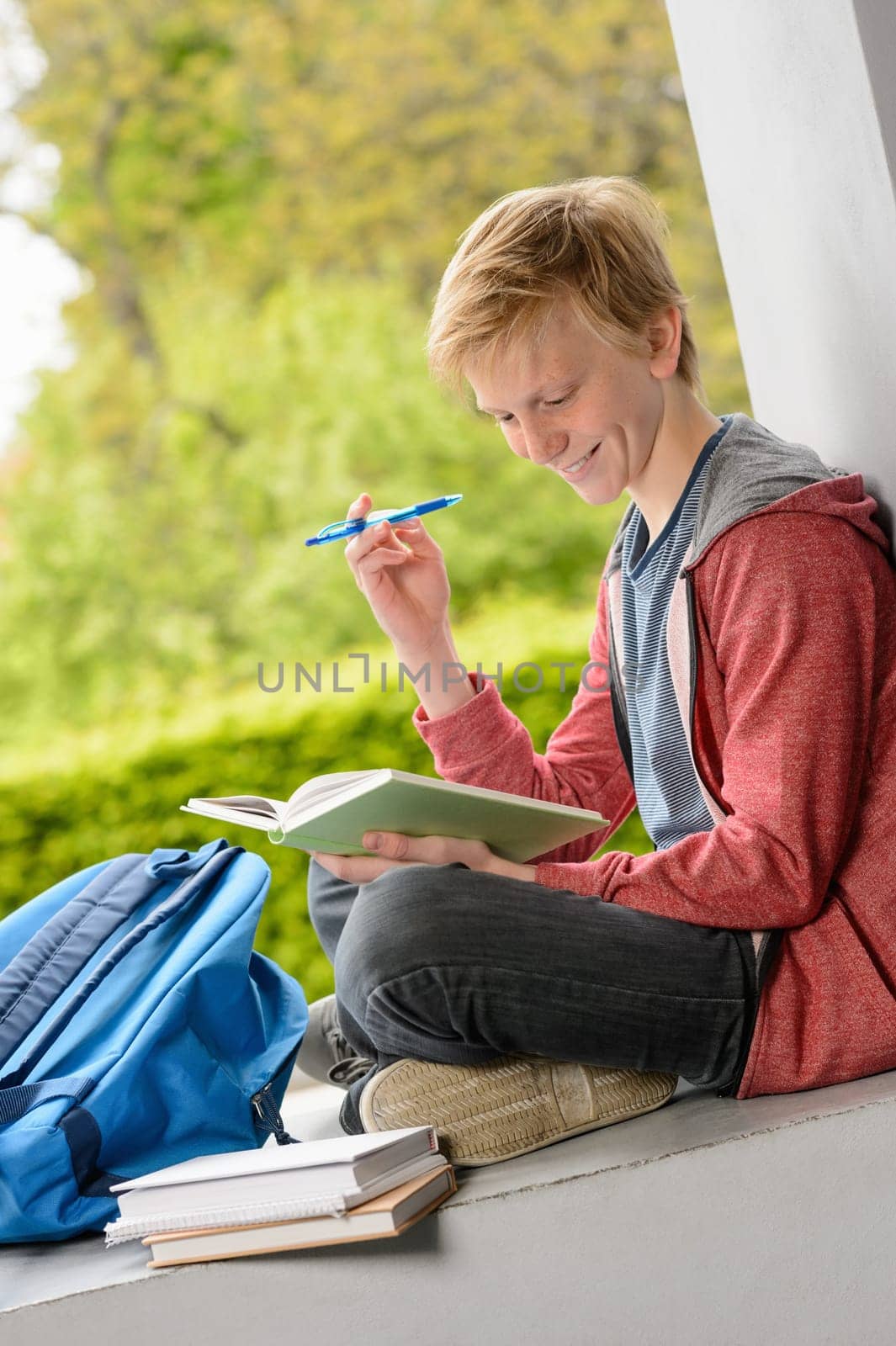 Smiling teenage boy reading book sitting on wall outside school