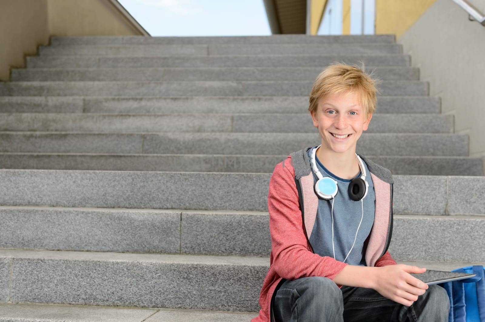 Happy teenage student boy with tablet sitting on steps outdoors