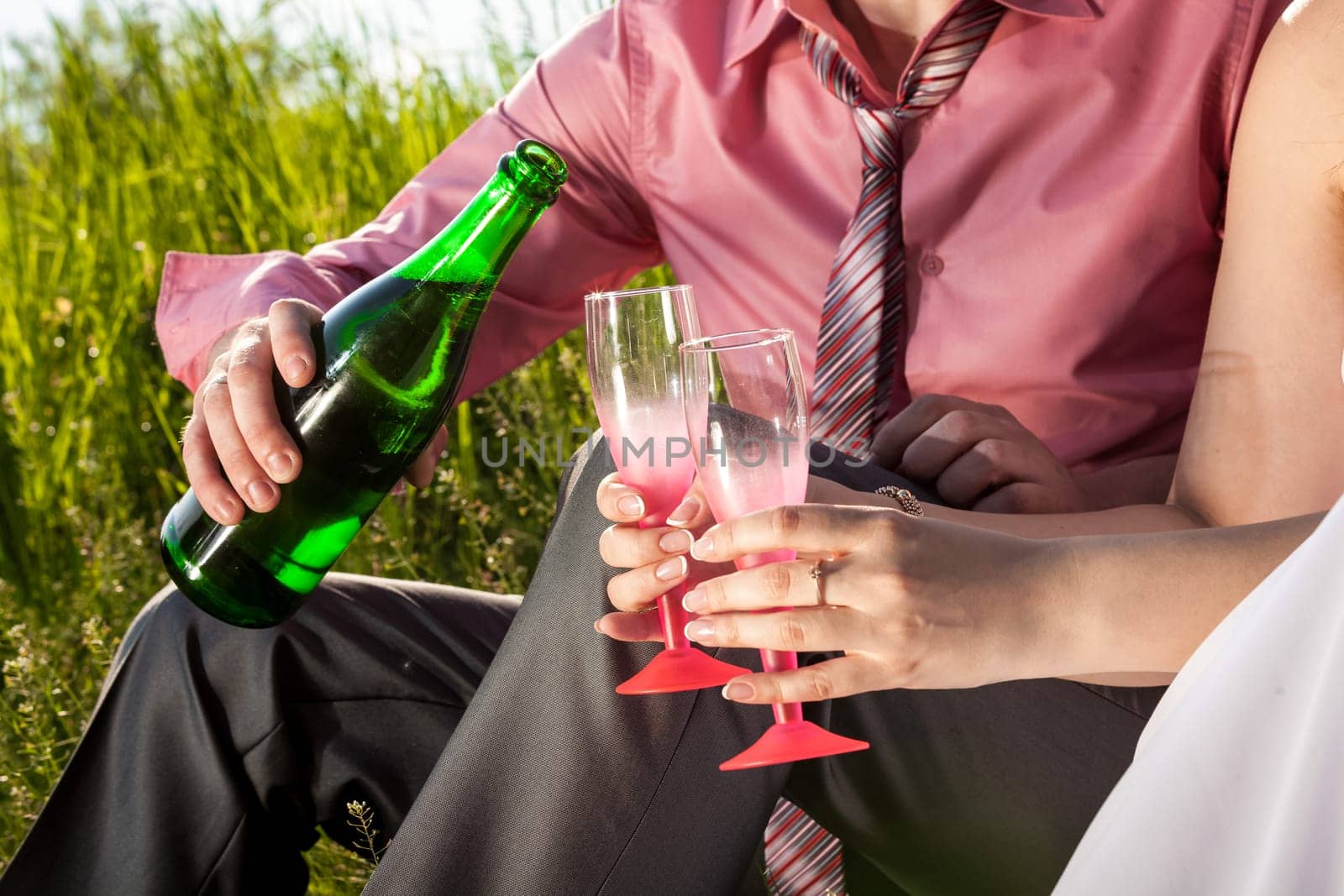 Closeup photo of groom pouring champagne at brides glass at meadow