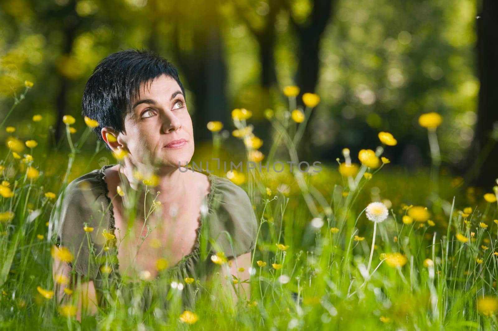 frontal head-and-shoulder view of a middle-aged black-haired woman on her belly in the grass with yellow flowers looking dreamily up the right