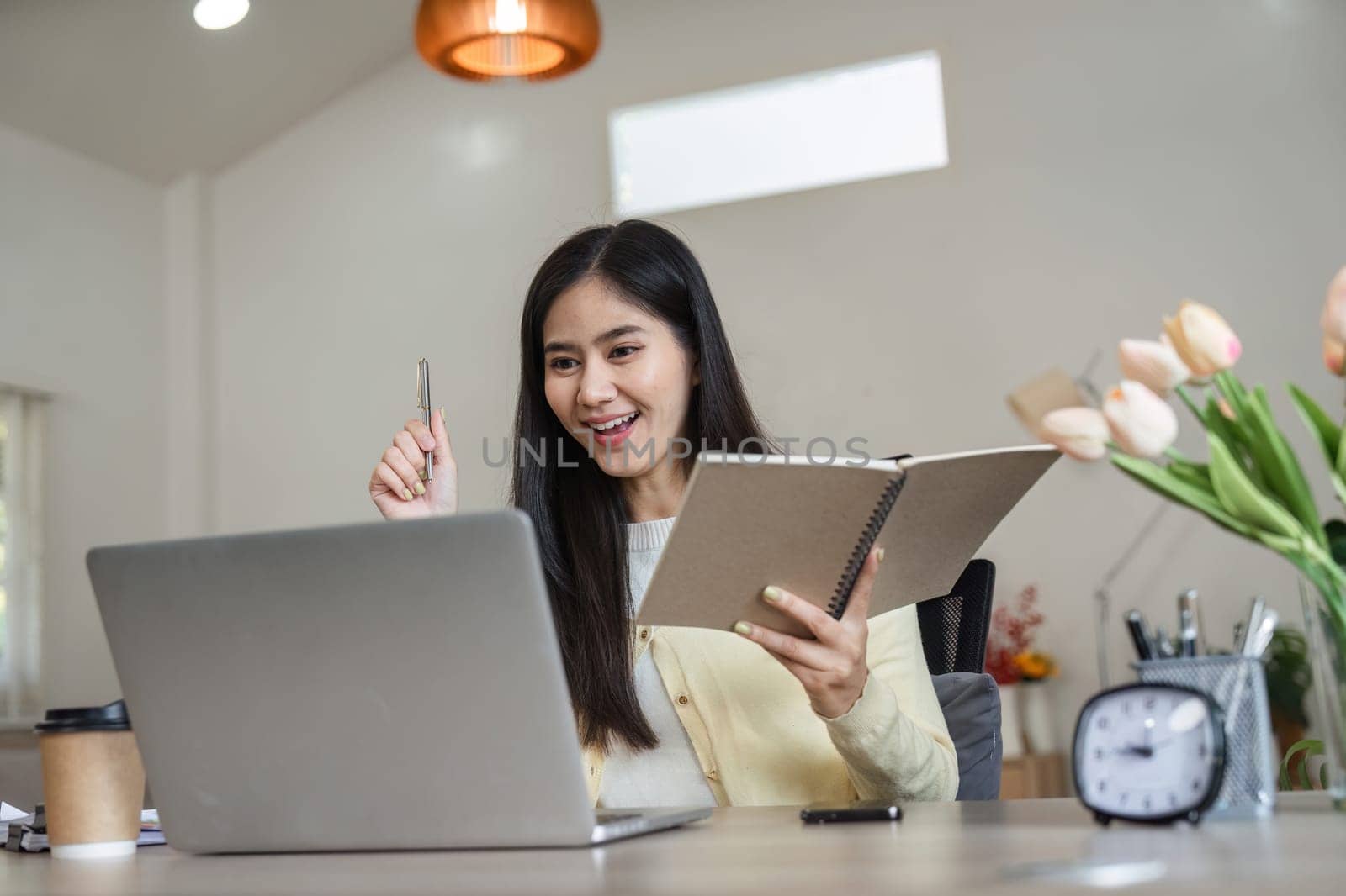 A joyful Asian woman holds a notebook and pen while smiling at her laptop, showcasing enthusiasm and engagement in her productive home office environment.