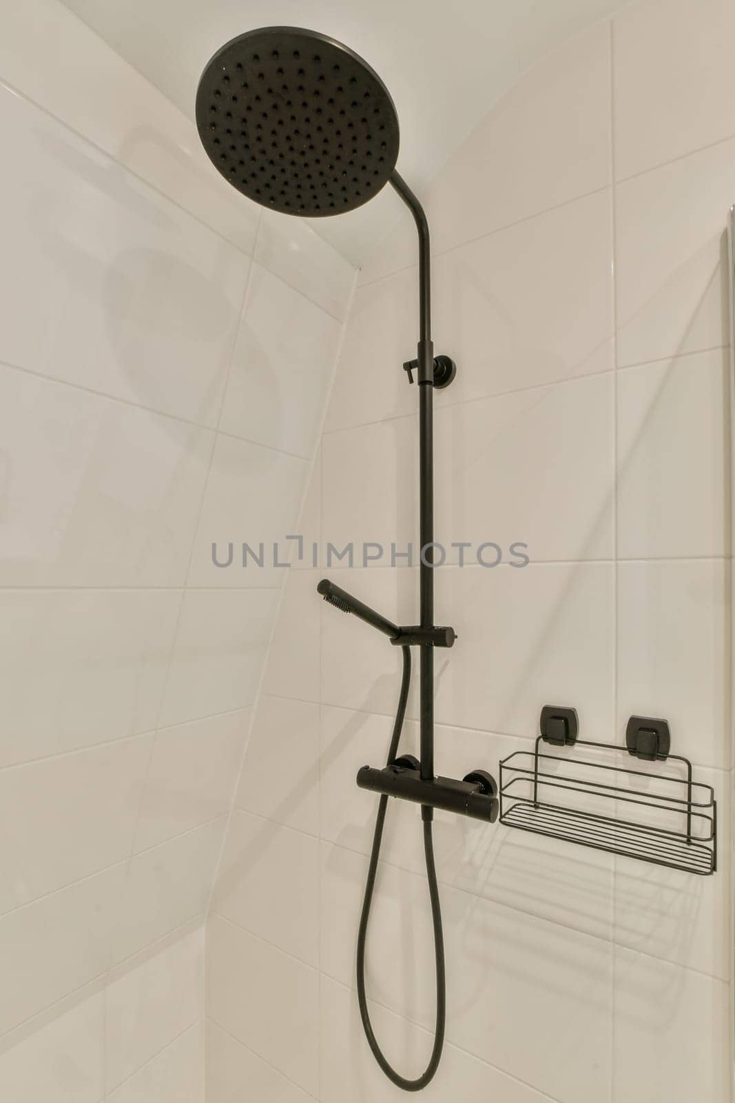 a black shower head in a white tiled bathroom with an overhead hand rail and wall mounted shower faucet