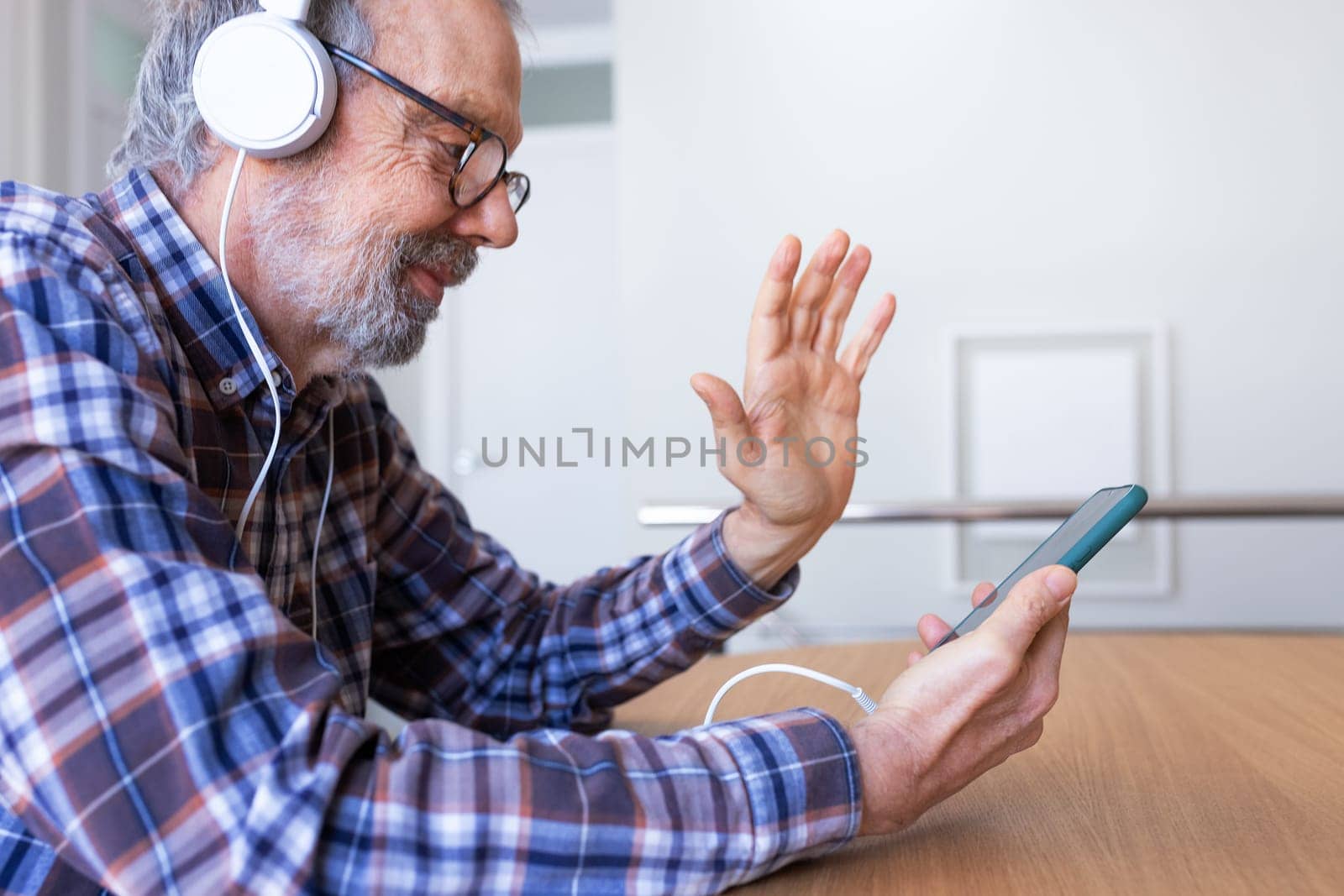 Side view of senior caucasian man waving hand during video call using phone and headphones by Hoverstock