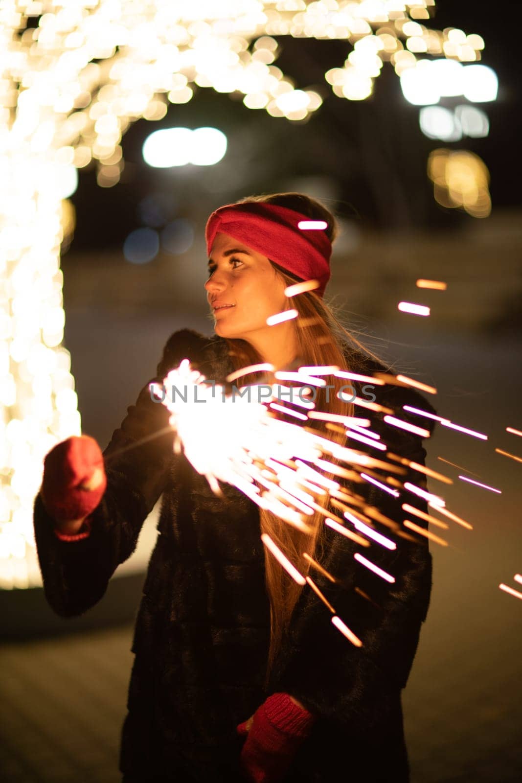 Woman holding sparkler night while celebrating Christmas outside. Dressed in a fur coat and a red headband. Blurred christmas decorations in the background. Selective focus by Matiunina