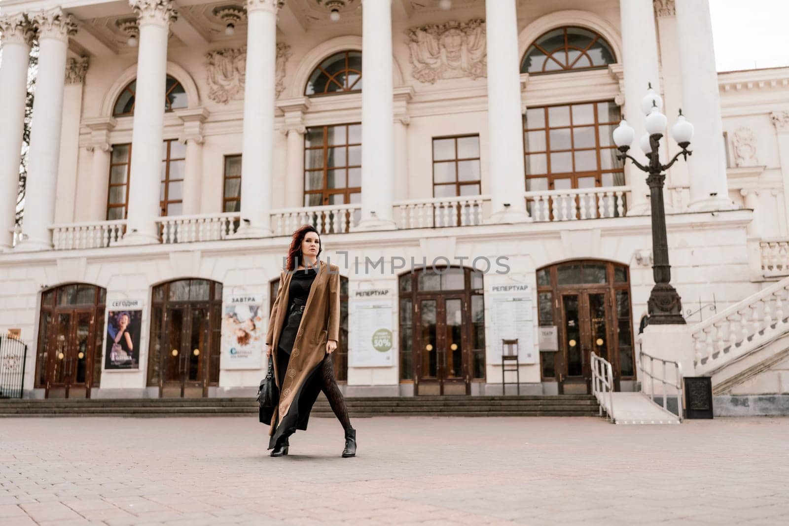 Woman street lifestyle. Image of stylish woman walking through European city on sunny day. Pretty woman with dark flowing hair, dressed in a beige raincoat and black, walks along the building. by Matiunina