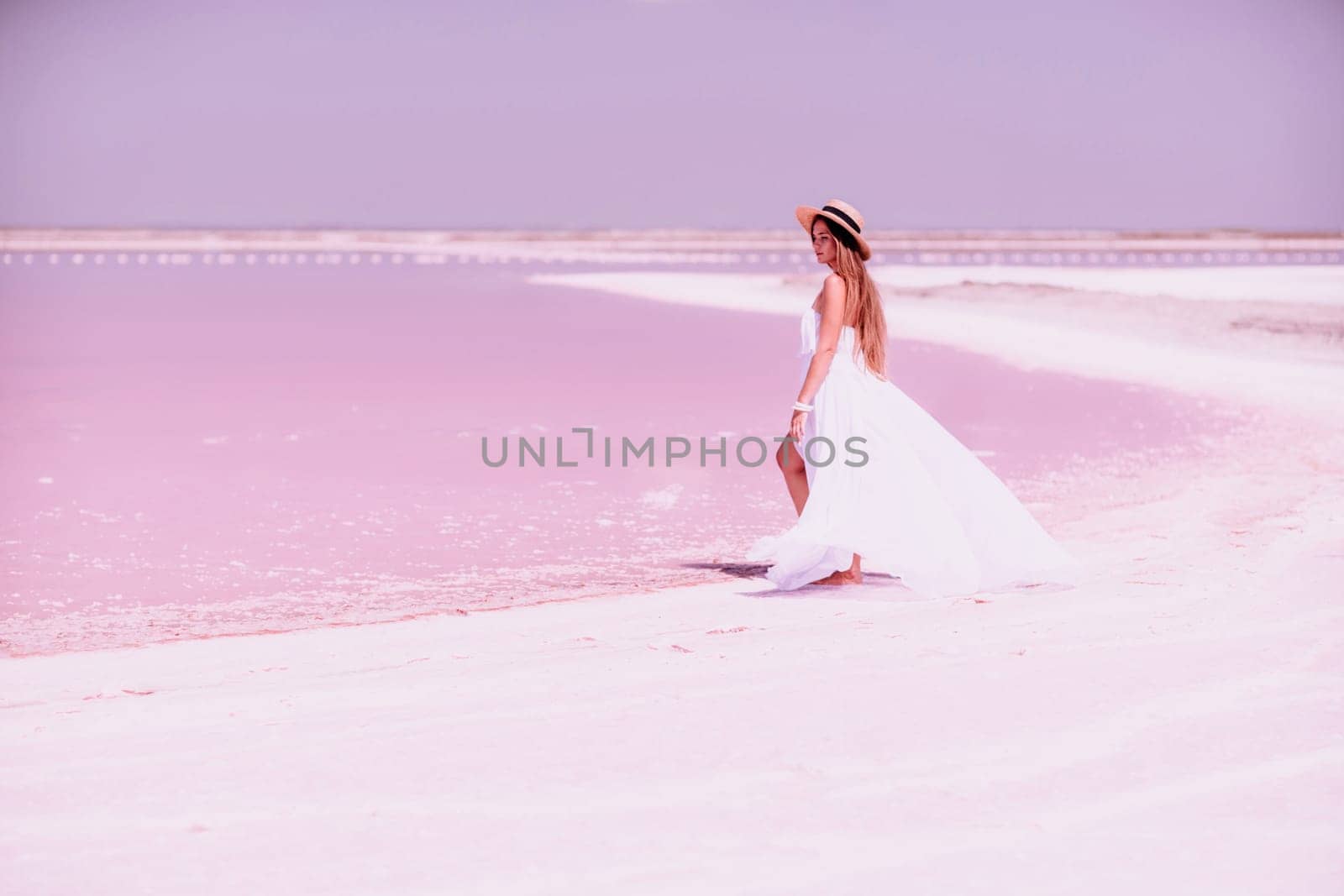 Woman in pink salt lake. She in a white dress and hat enjoys the scenic view of a pink salt lake as she walks along the white, salty shore, creating a lasting memory. by Matiunina