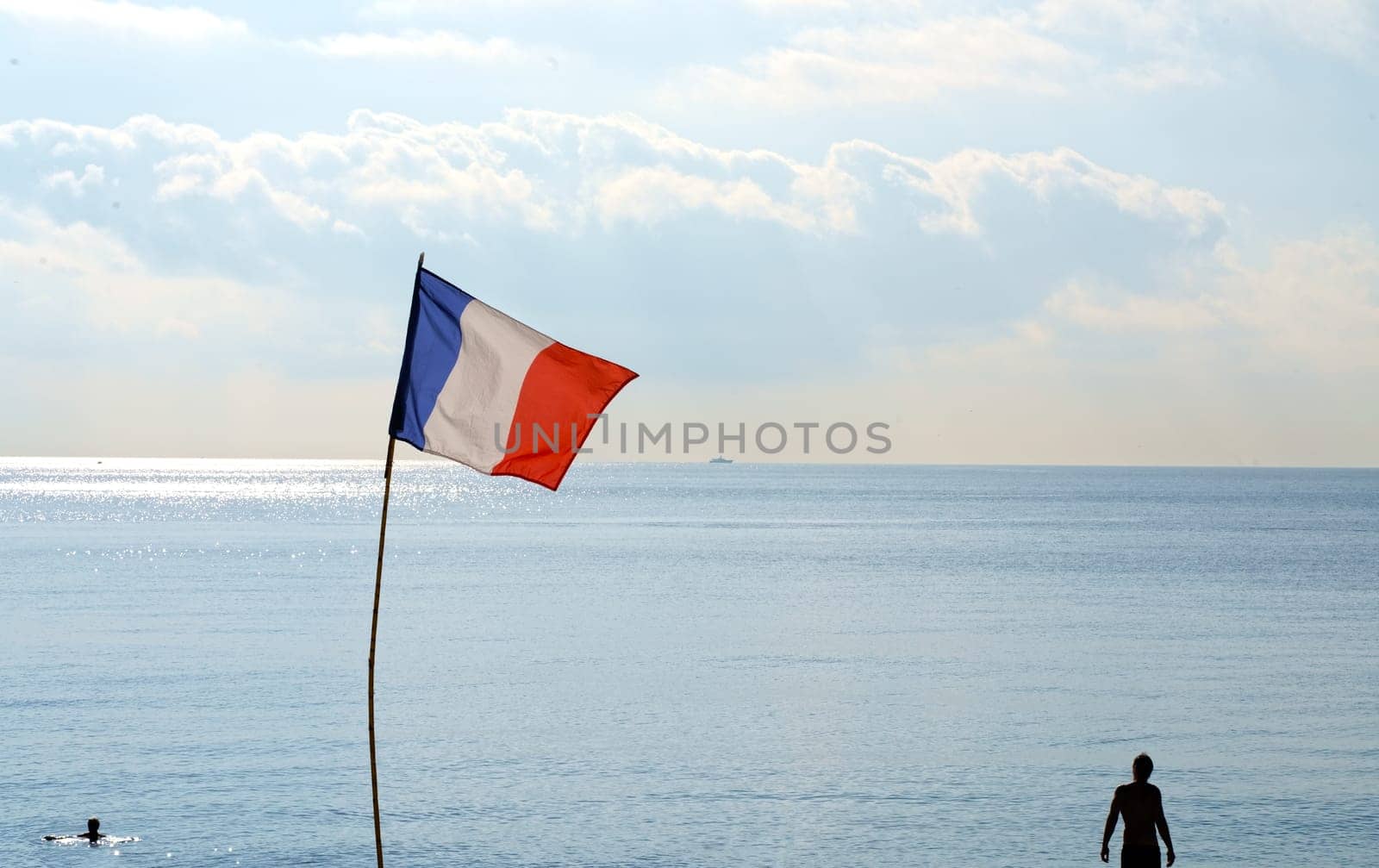 French flag. The French flag flutters against the backdrop of the summer sea and blue cloudless sky. The atmosphere of a picturesque beach. People sunbathe and swim in the sea by aprilphoto