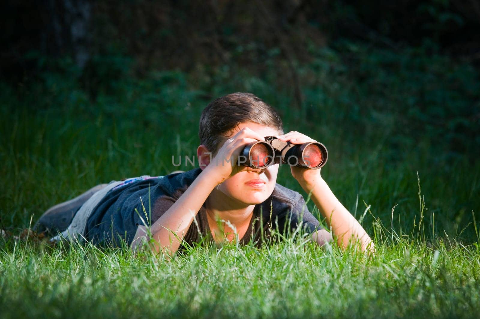 Half-side full-body view of a 13-year-old male teenager lying on his stomach in the woods and looking through binoculars