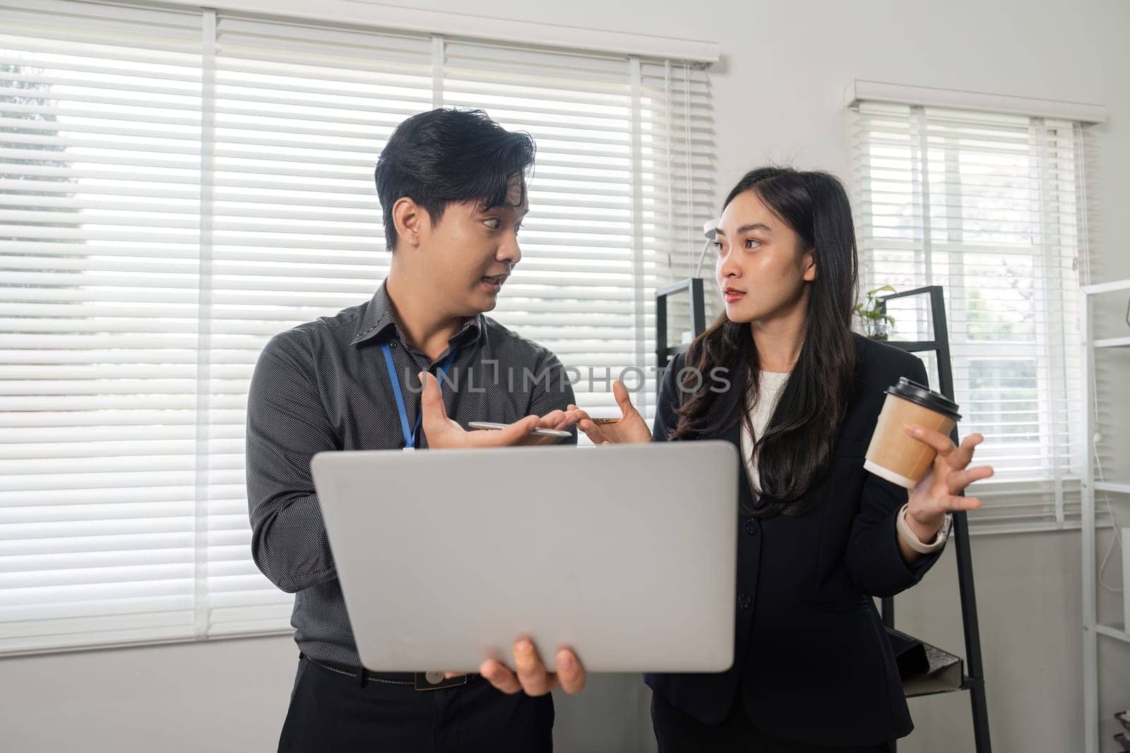 Two business professionals conversing and reviewing information on a laptop while one holds a coffee cup in a collaborative office setting.