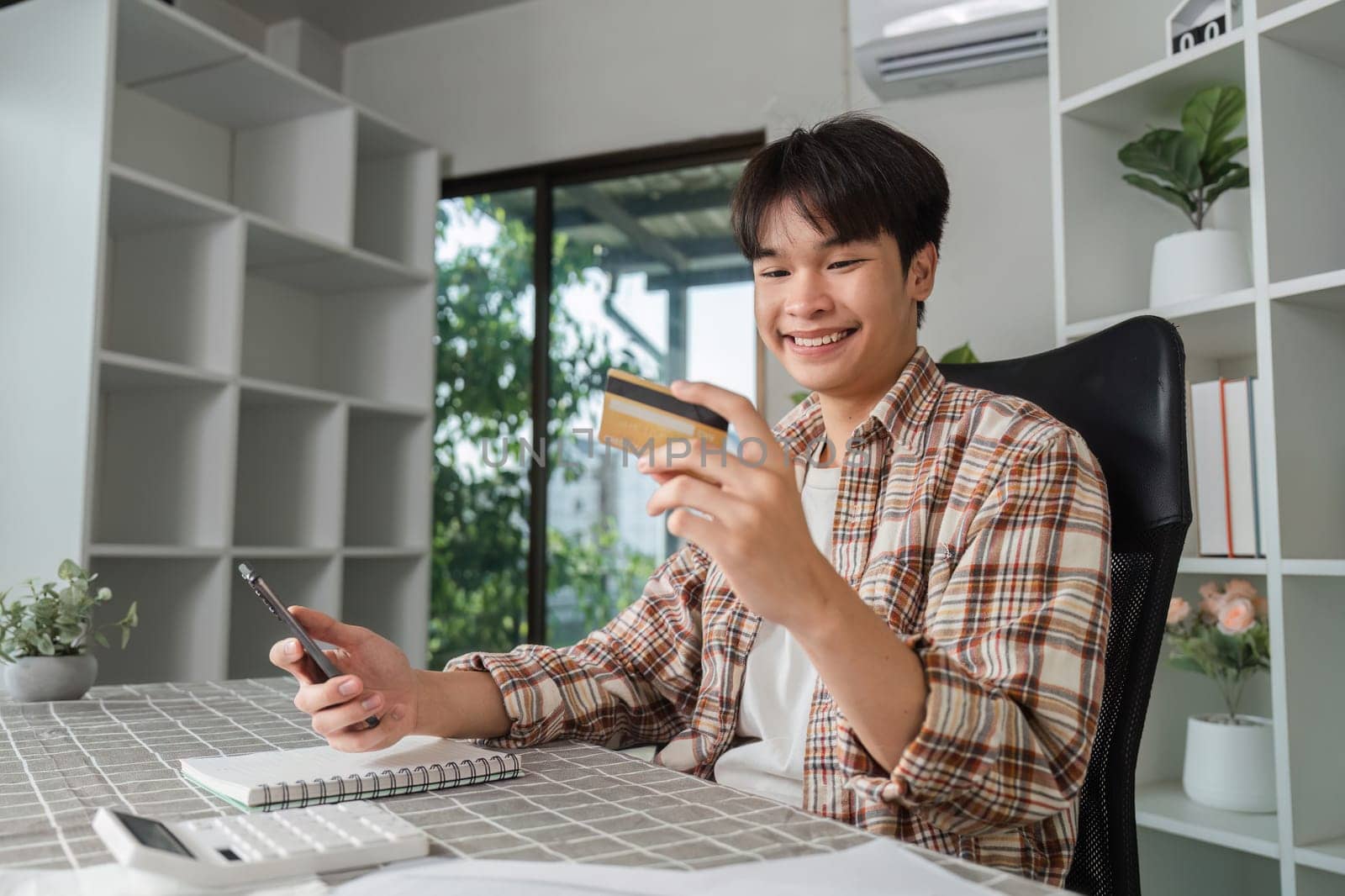 A joyful young man holds a credit card and smartphone, celebrating financial success in his home office, representing positive finance management.