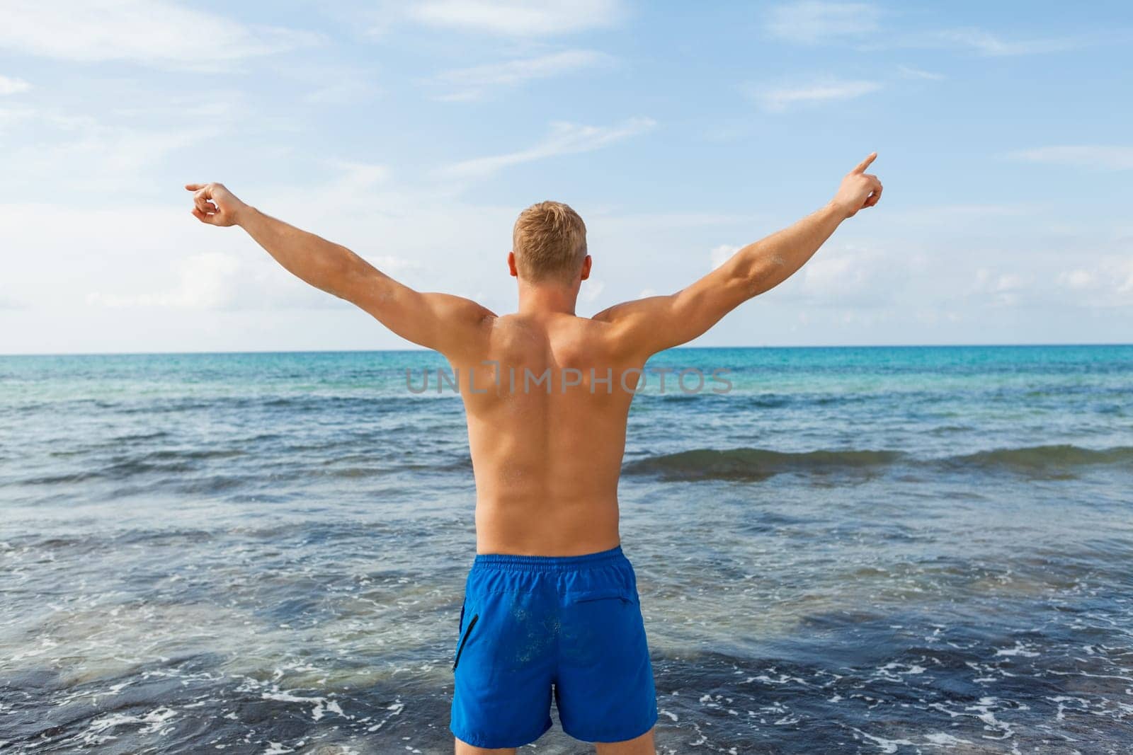 Young man in blue swimming trunks stands on the beach in the blue sea on holiday