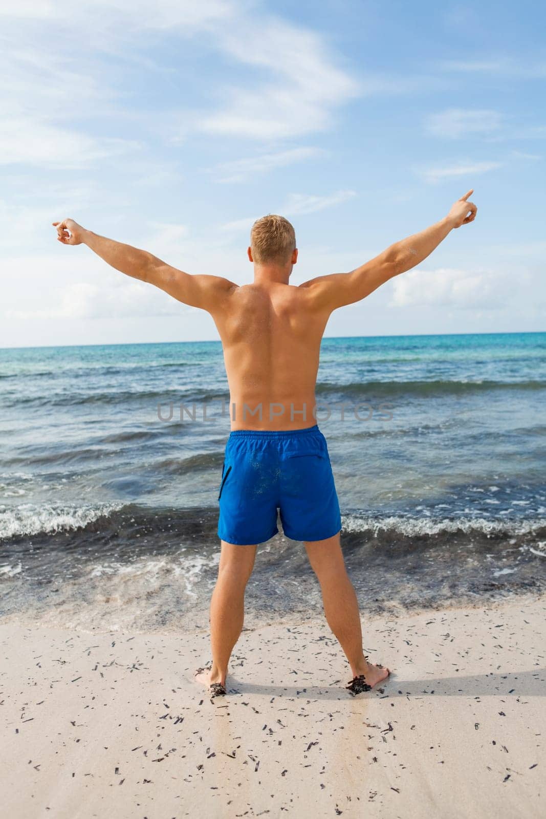 Young man in blue swimming trunks stands on the beach in the blue sea on holiday