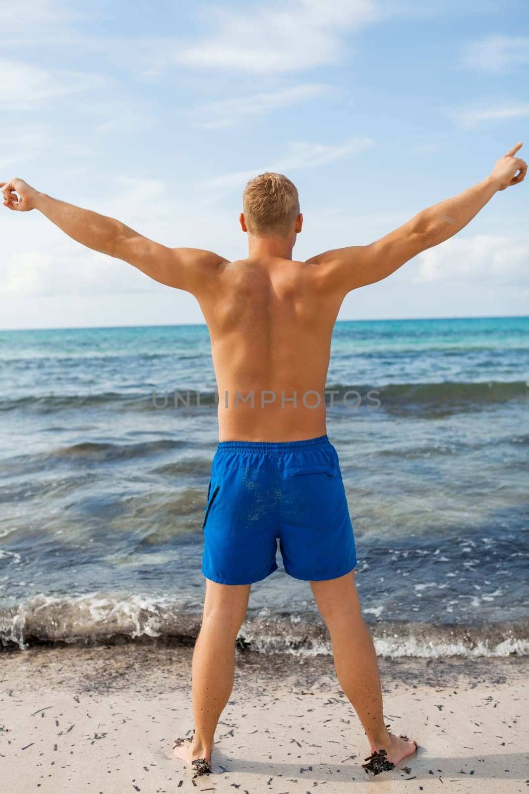 Young man in blue swimming trunks stands on the beach in the blue sea on holiday