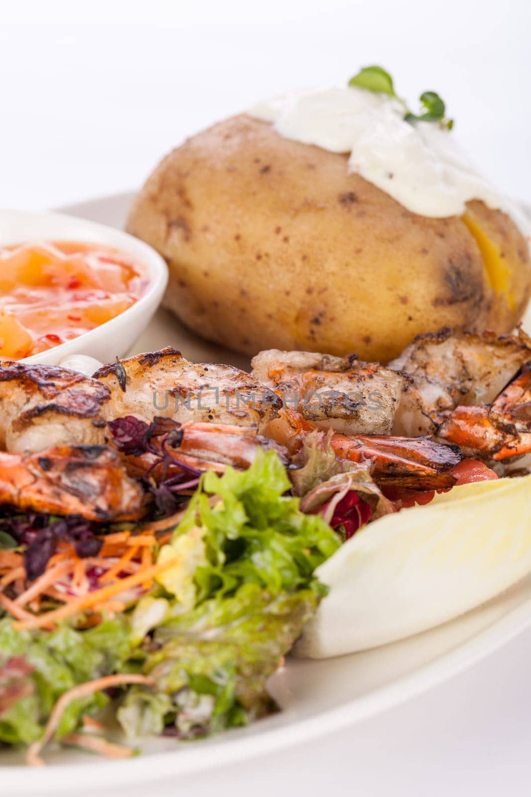 Roasted shrimp on a skewer on mixed salad with dip and foil potato prepared on a plate insulated against a white background