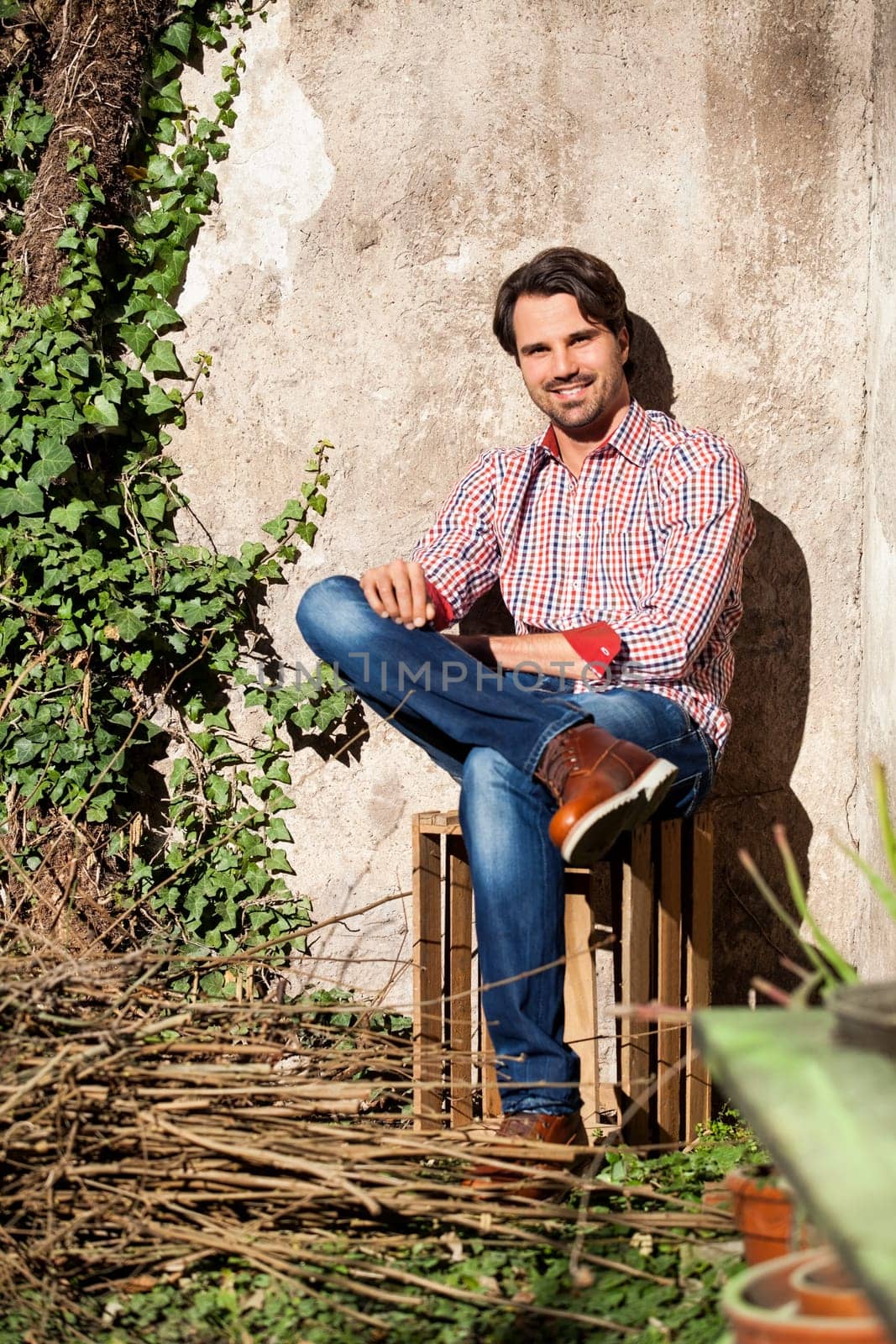 Young man with a plaid shirt and jeans with dark hair and beard outdoors under an efeuranke in daylight
