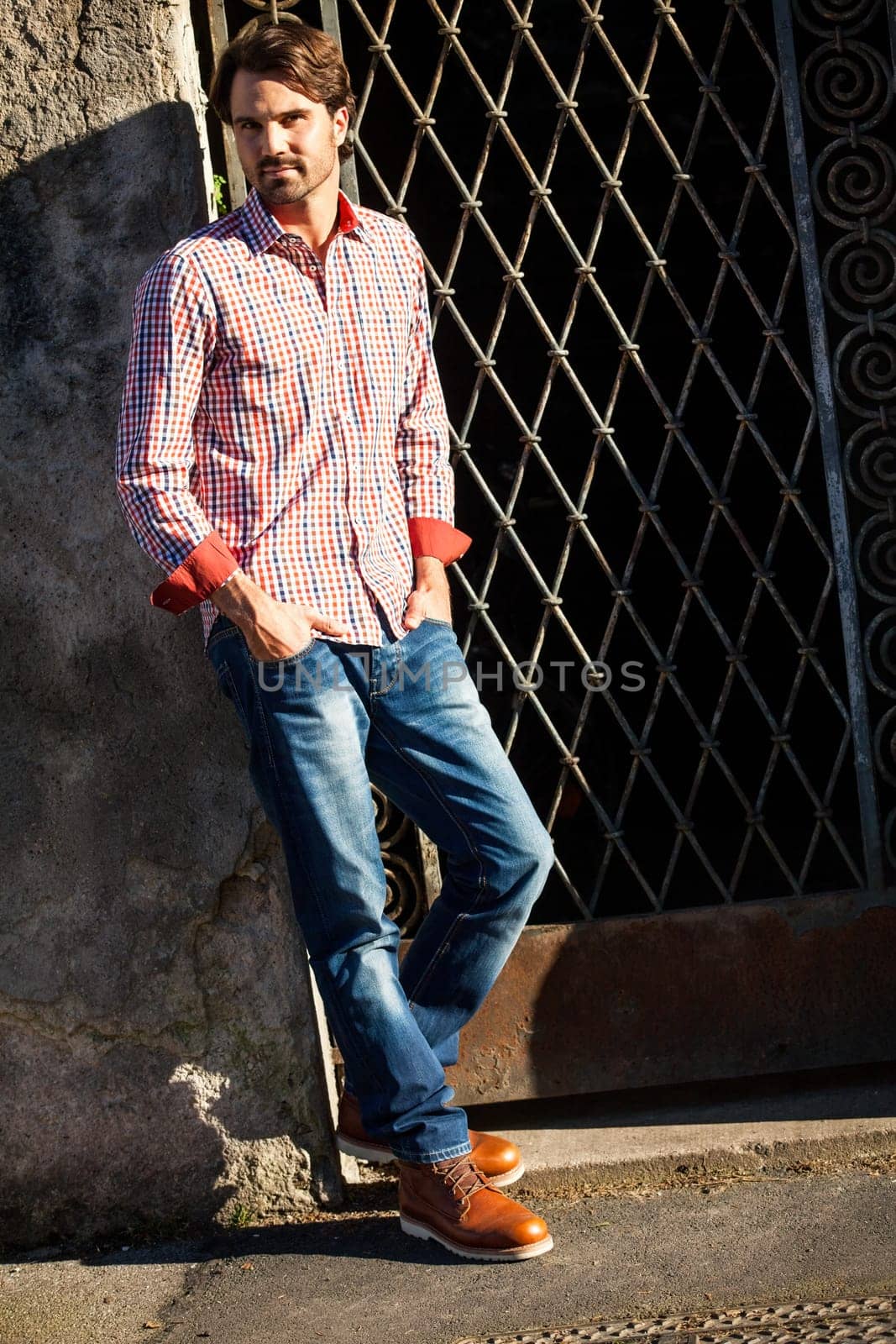 Attractive young man with a plaid shirt and jeans leaning against a wall outdoors in summer