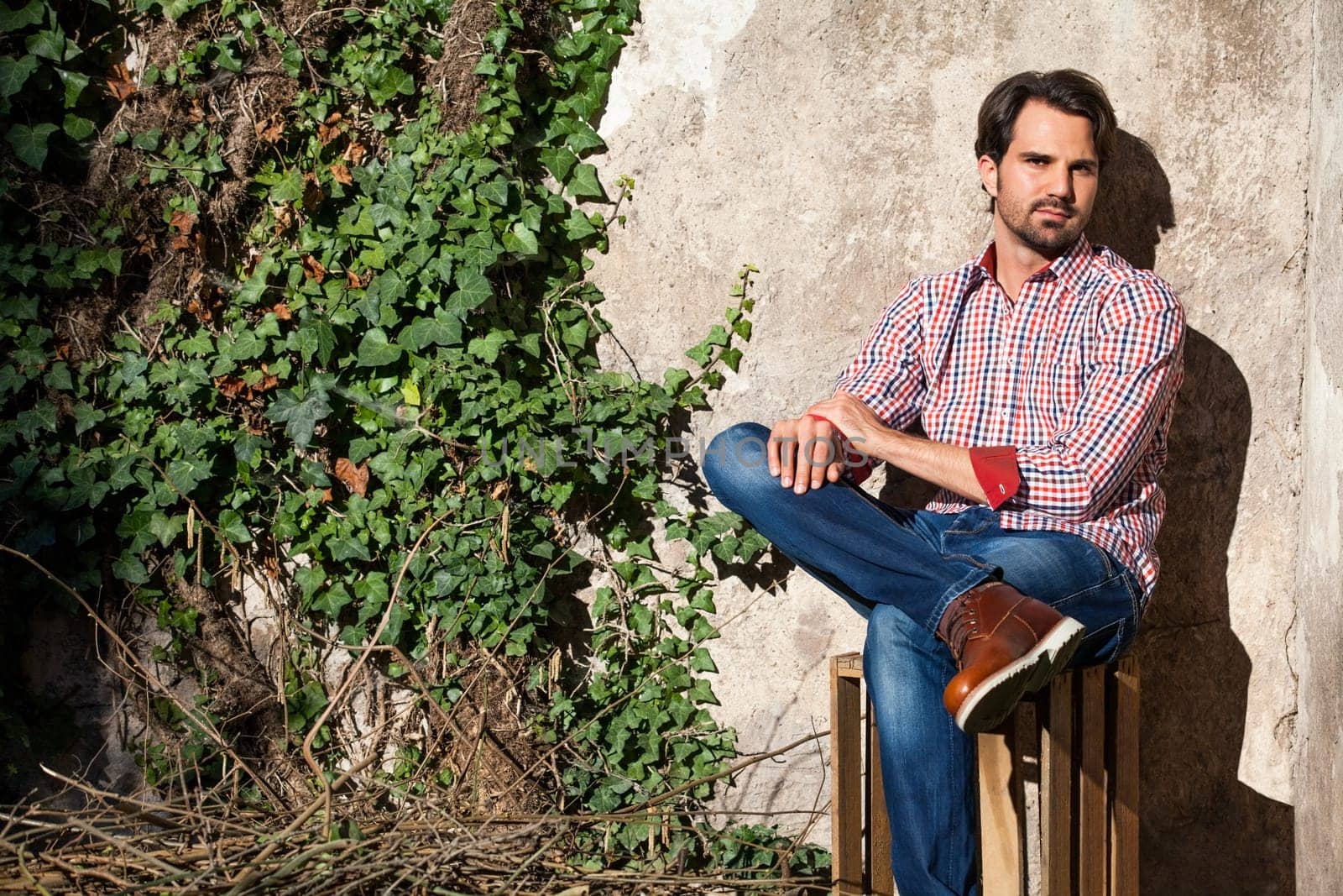 Young man with a plaid shirt and jeans with dark hair and beard outdoors under an efeuranke in daylight