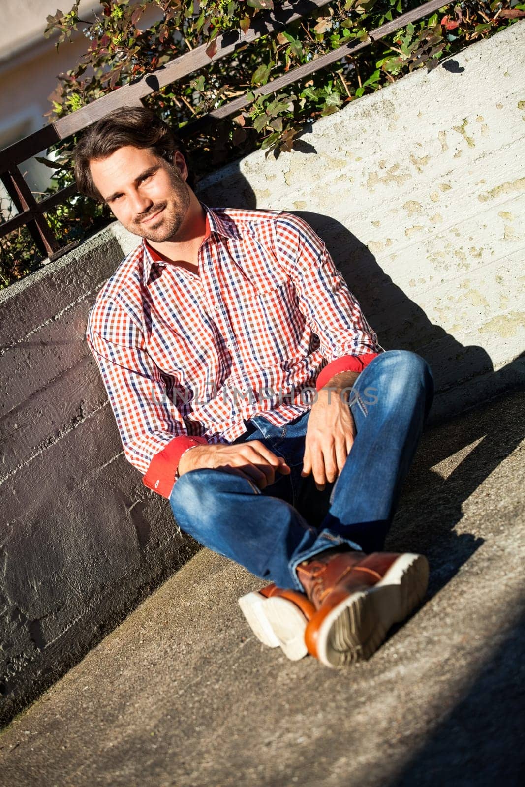 Young man with a checked shirt sits on a wall in the sun and relaxes