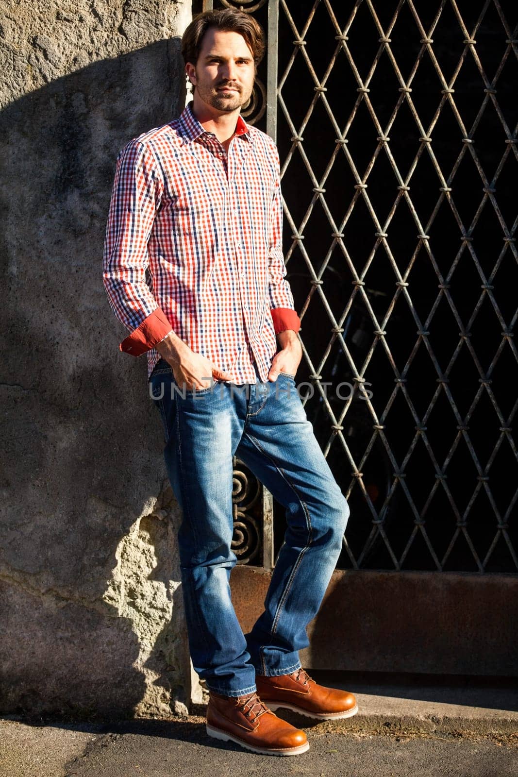 Attractive young man with a plaid shirt and jeans leaning against a wall outdoors in summer