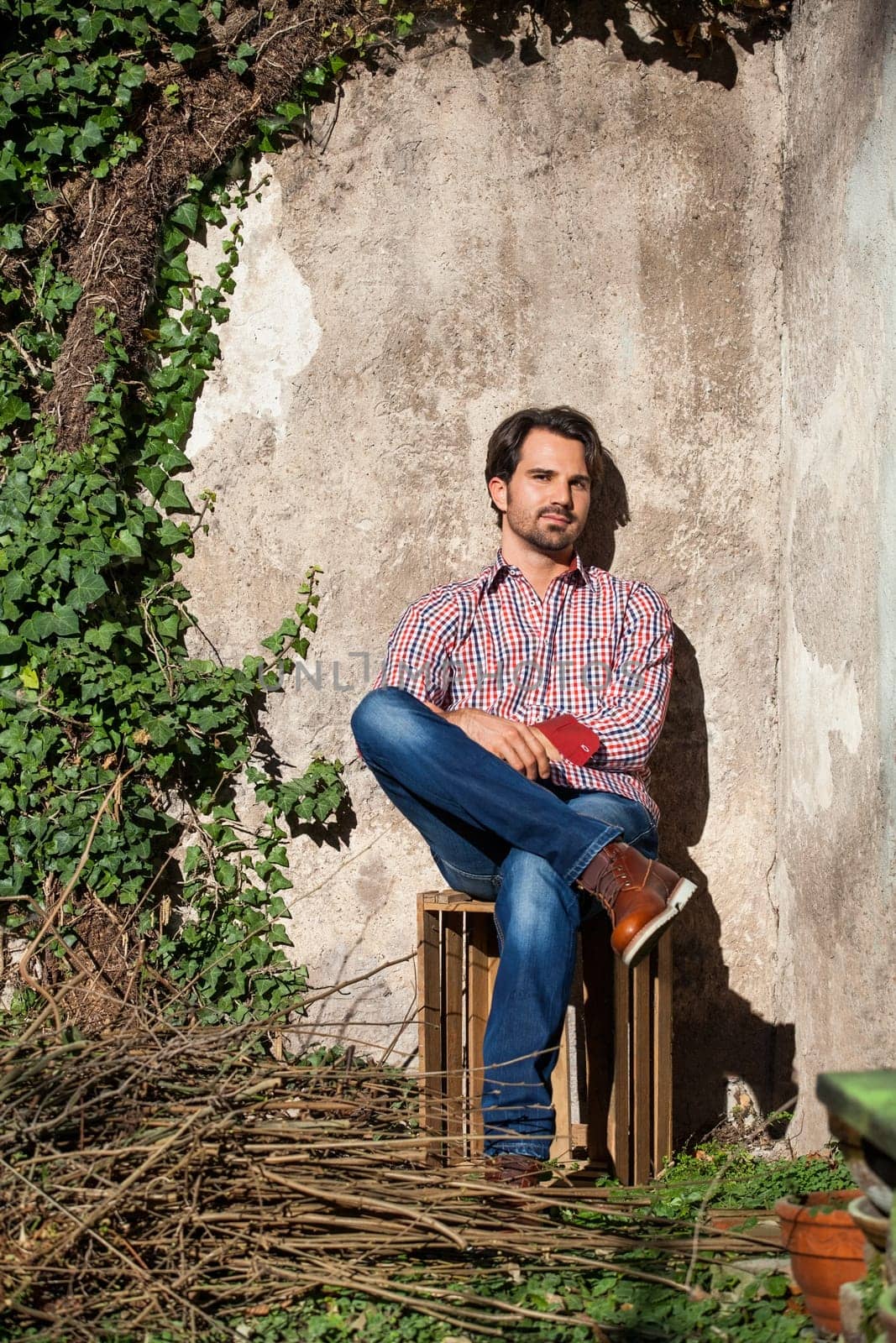 Young man with a plaid shirt and jeans with dark hair and beard outdoors under an efeuranke in daylight