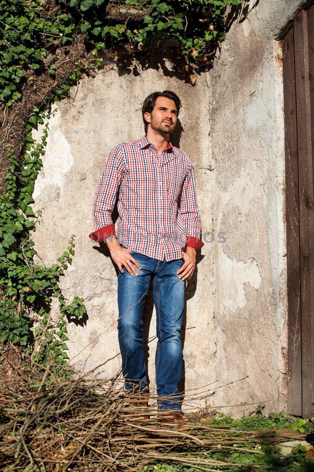 Young man with a plaid shirt and jeans with dark hair and beard outdoors under an efeuranke in daylight
