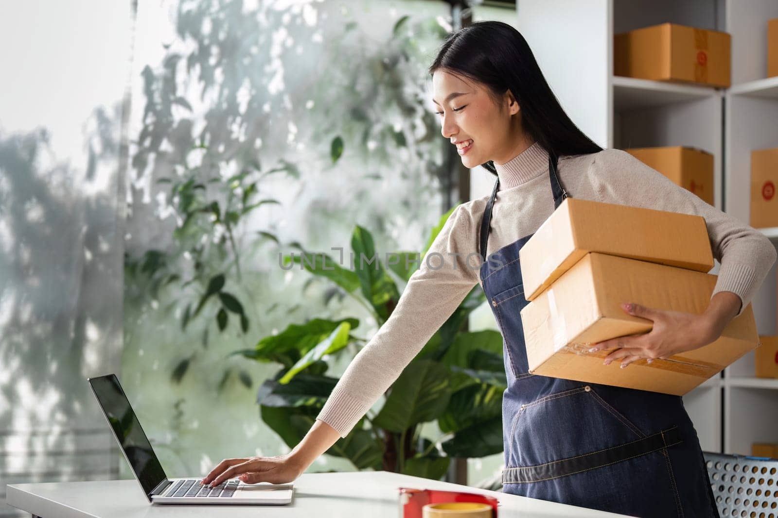 A young woman in an apron carries two boxes while working on a laptop in an organized workspace filled with greenery.
