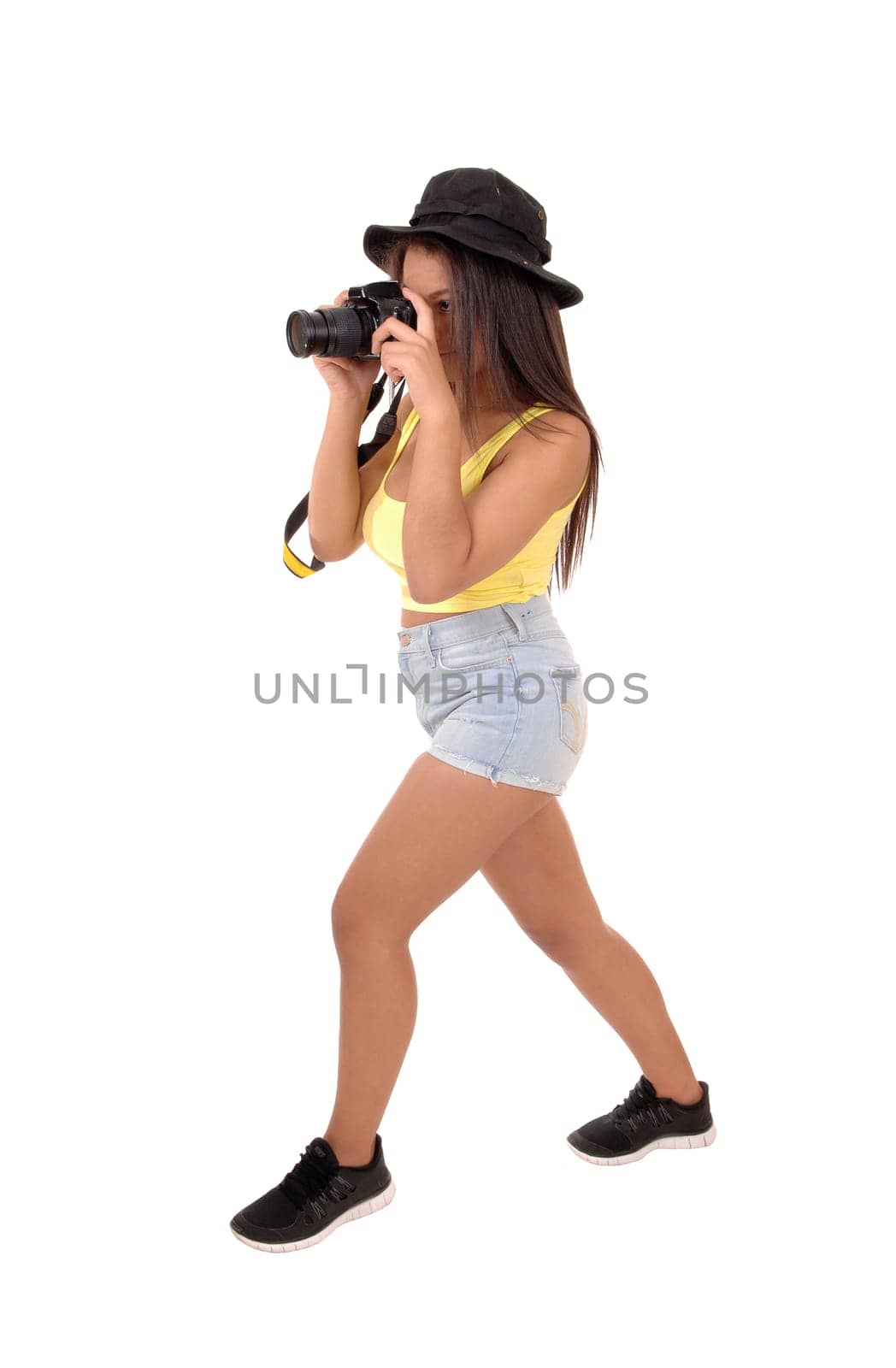 A young woman in shorts and a hat standing in the studio and taking.pictures with her camera, isolated over white background..