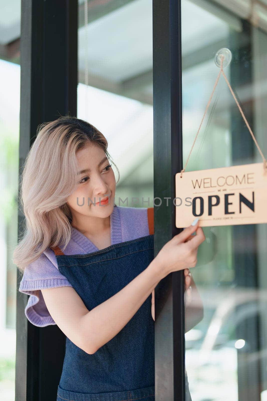 open a small business Portrait of happy Asian woman wearing apron holding welcome we are open sign at front of cafe. by Manastrong