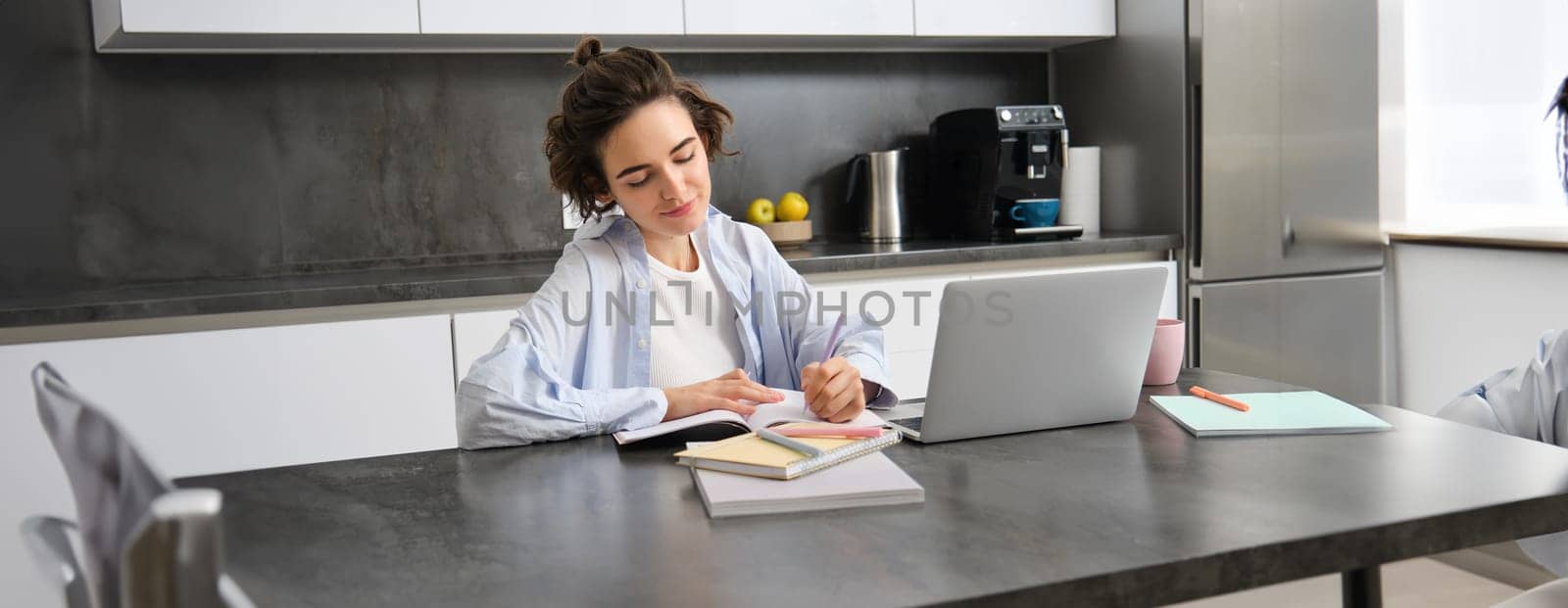 Image of young woman managing her bills, works from home with laptop, writes down information on paper. Student does homework in her kitchen.