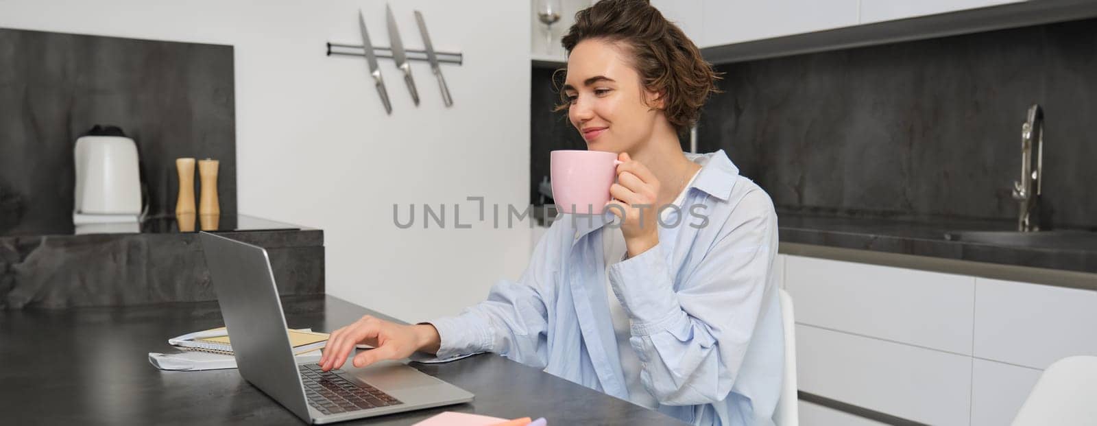 Beautiful girl working from home, drinking coffee and looking at laptop. Young businesswoman sits in kitchen and manages her business via computer.