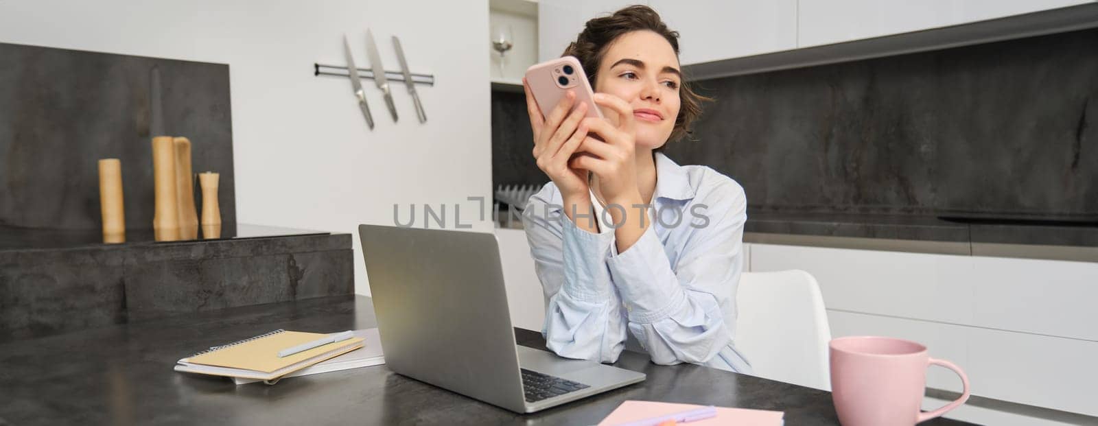 Portrait of brunette woman chats on mobile phone, uses smartphone app from home, orders delivery while sits in kitchen by Benzoix