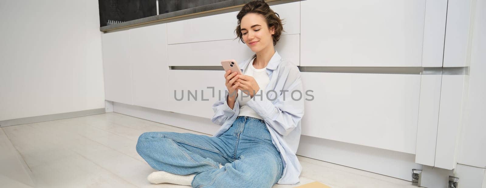 Portrait of girl with mobile phone, sits on floor at home, orders takeaway in app. Young woman with smartphone does online shopping from home, browsing websites by Benzoix