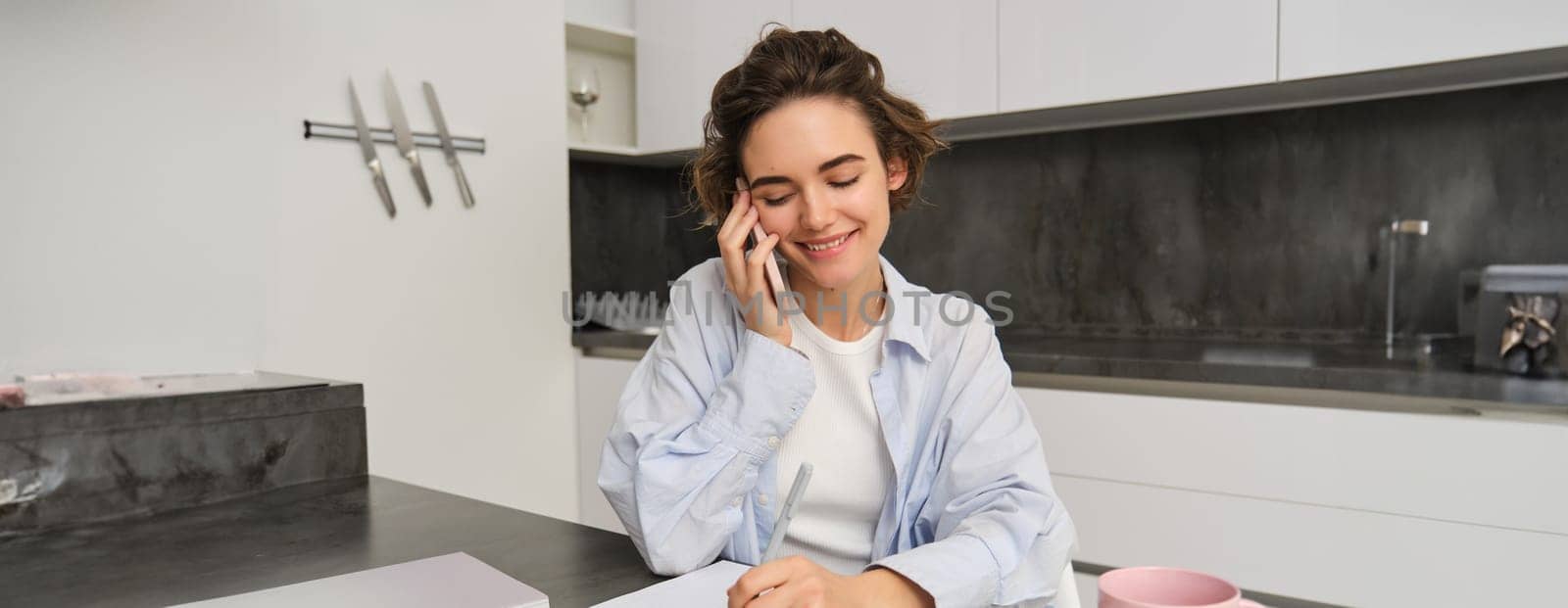 Image of woman working from home, writing down information during a phone call, calling someone, using smartphone.