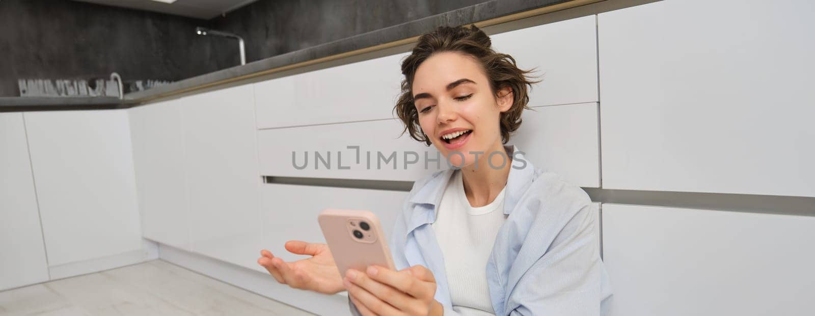 Portrait of happy young woman, shows her brand new kitchen, sits on floor, points at furniture and smiles, video chats on smartphone. Technology and lifestyle concept