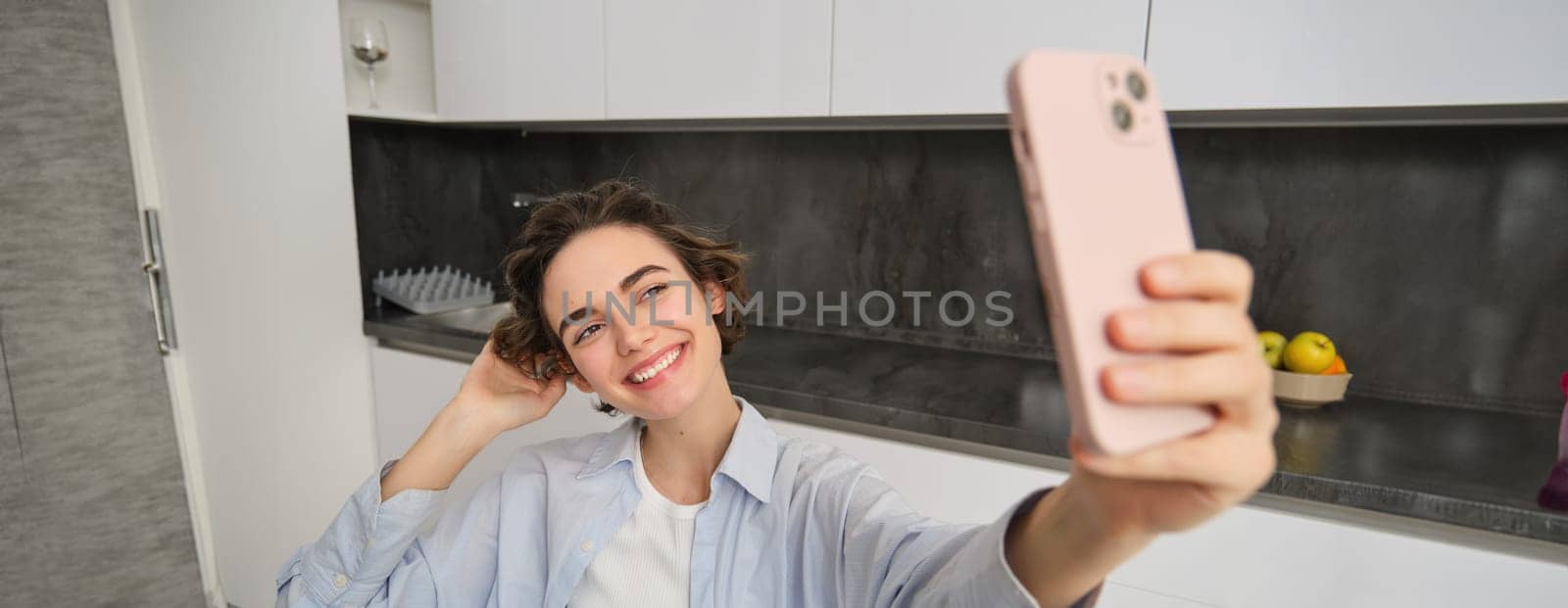 Portrait of beautiful young woman, taking selfie on mobile phone at home, poses for photo with smartphone.