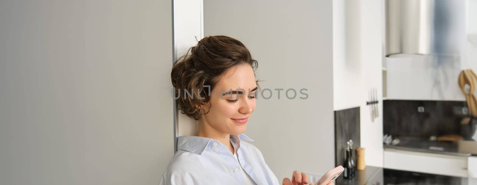 Portrait of smiling, beautiful woman, looking at smartphone, standing at home near kitchen, checking messages, chatting on mobile phone by Benzoix