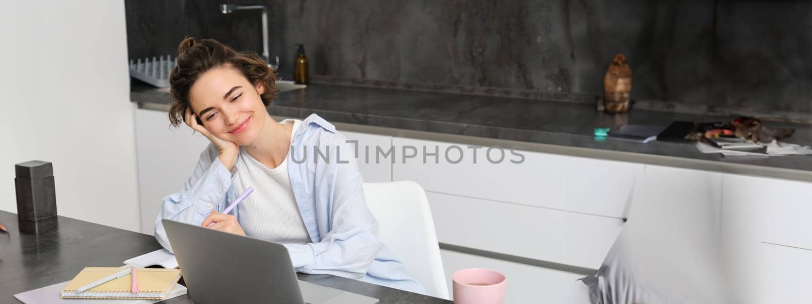 Image of young woman works on laptop at home, productive girl studies in her kitchen on computer, listens to webinar course by Benzoix