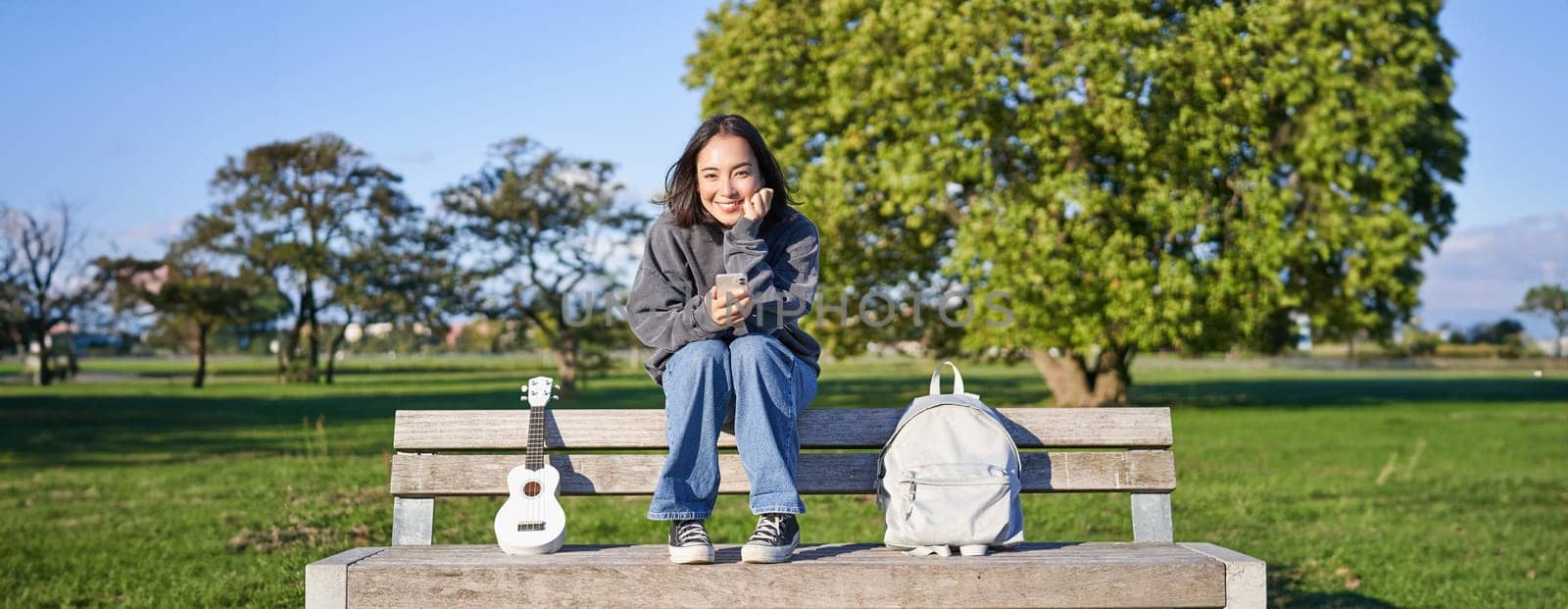 Beautiful brunette girl on bench in park, sitting with ukulele and backpack, holding smartphone, using mobile app by Benzoix