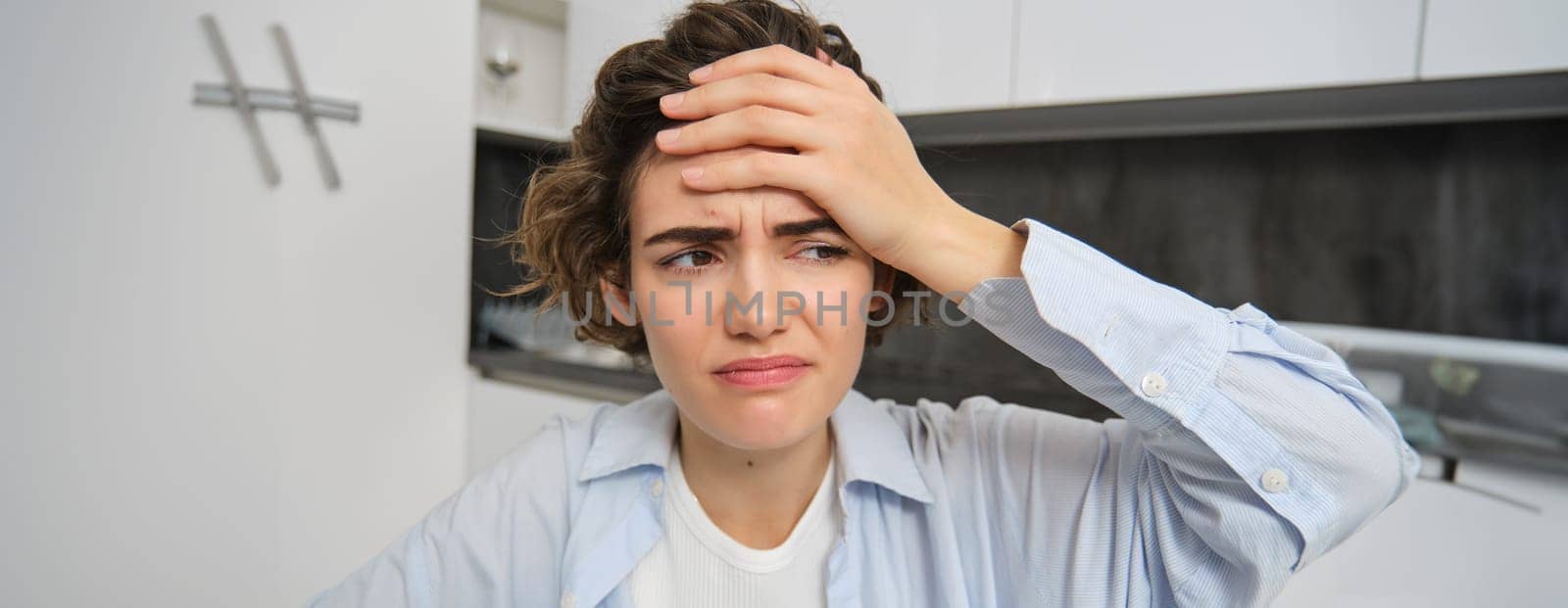 Portrait of woman with mgiraine. Girl grimaces from painful headache, touches her head, frowns from discomfort, sits at home.