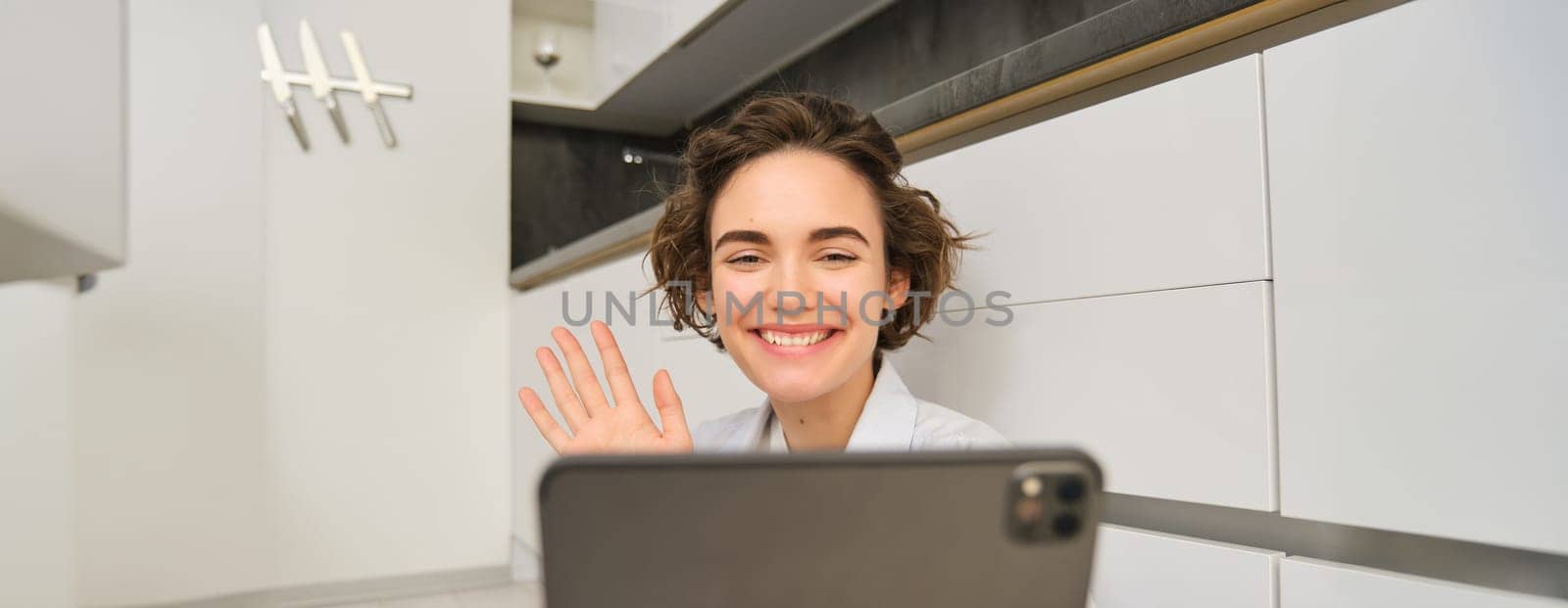 Close up portrait of brunette girl, smiles and looks at tablet, waves at camera, connects to online meeting, chats with friends, sits on floor at home in kitchen.