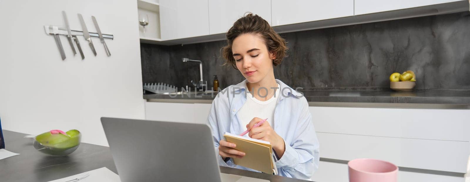 Working woman makes notes, writes down information on notebook, sitting with laptop at home.