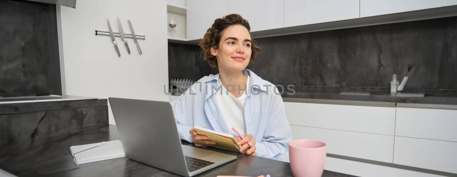 Image of young woman, entrepreneur works from home. Girl studies remote, writes down information, makes notes, uses laptop, does homework online.